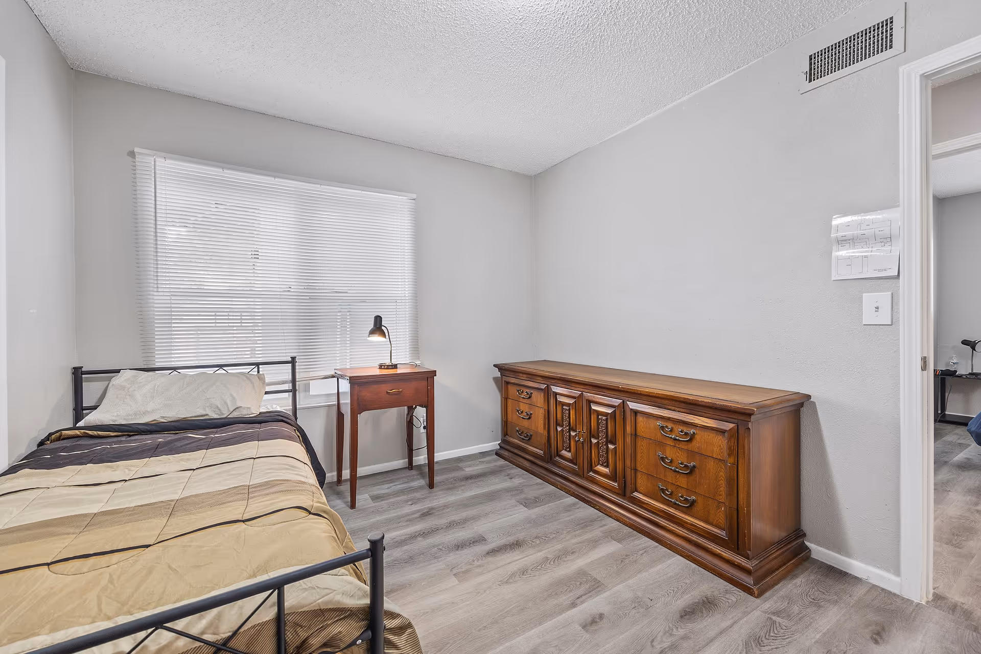 Small bedroom with a single bed, a bedside table and lamp, and a long wooden dresser against a light gray wall.