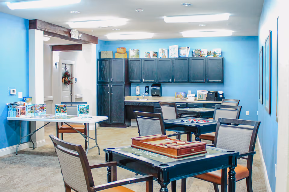 A bright activity room with blue walls featuring several tables and chairs arranged for games and puzzles. One table has a backgammon set, another has a checkerboard, and a folding table holds several puzzle boxes. Dark wood cabinets and a countertop with a sink and dispensers are along the back wall. The room is well-lit with ceiling lights.