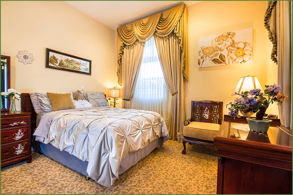 Cozy, traditionally furnished bedroom with a made bed, ornate drapery over a window, wooden nightstands and floral decorations.