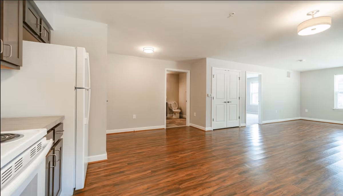 Interior view of an unfurnished apartment with wood flooring, white walls, and ceiling lights. The image shows a kitchen area with white appliances and dark cabinets on the left, an open living space, a doorway leading to a bathroom with a visible toilet, and another doorway leading to a separate room with a window.