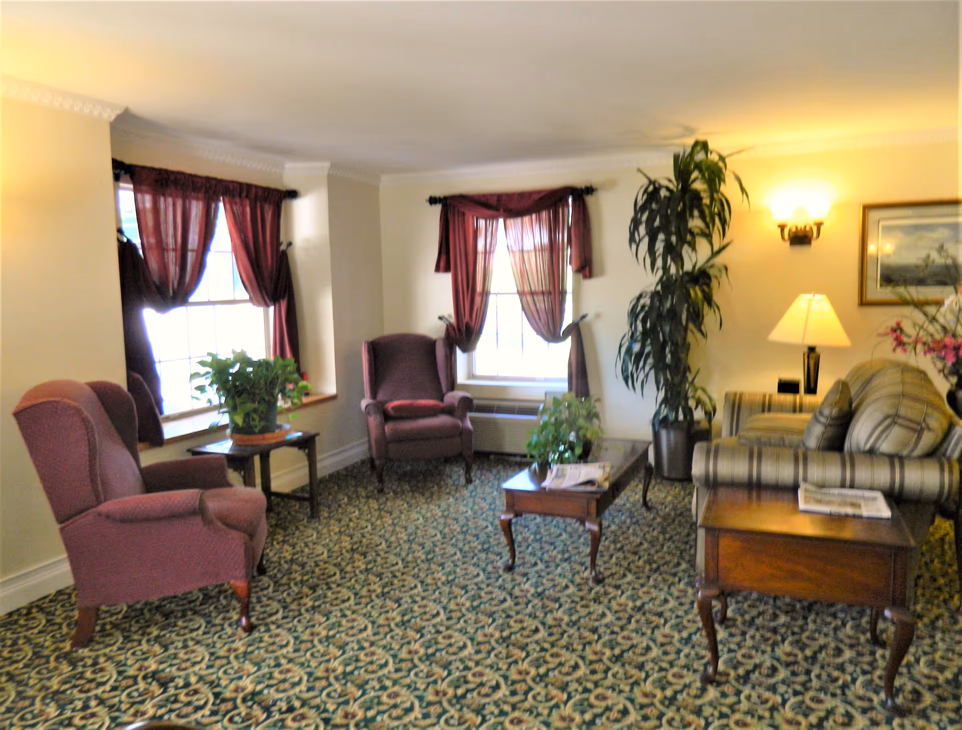 A cozy living room with patterned carpet, two burgundy armchairs, a striped sofa, wooden side tables, a coffee table with plants and newspapers, burgundy curtains on two windows, a tall potted plant, a table lamp, and a wall sconce light.