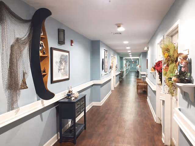 Long decorated indoor hallway with light blue walls, wood flooring, framed artwork and small tables in an assisted living facility.