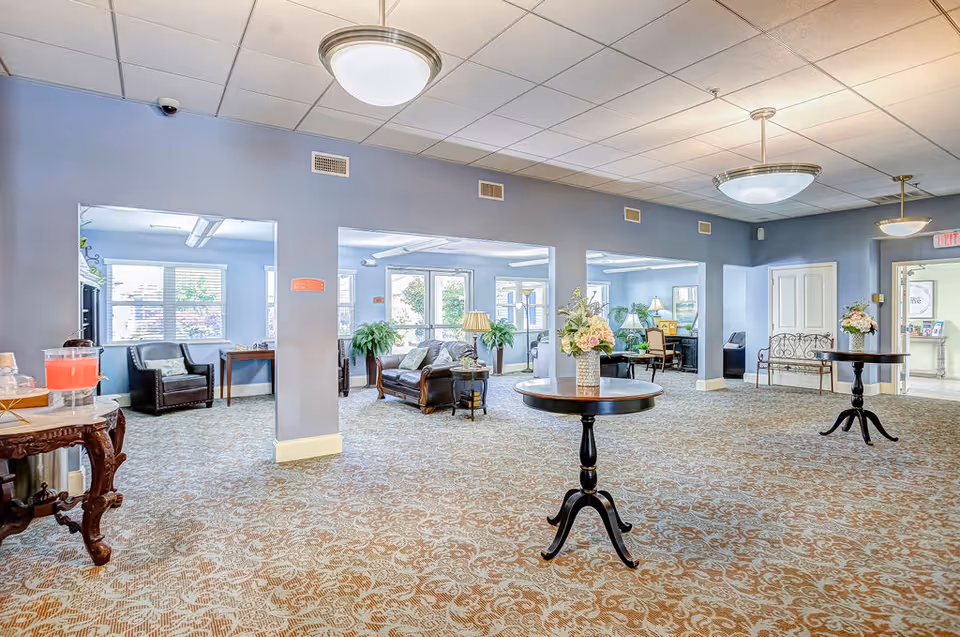 Spacious senior living lounge with seating areas, round tables topped with floral arrangements, and soft overhead lighting.