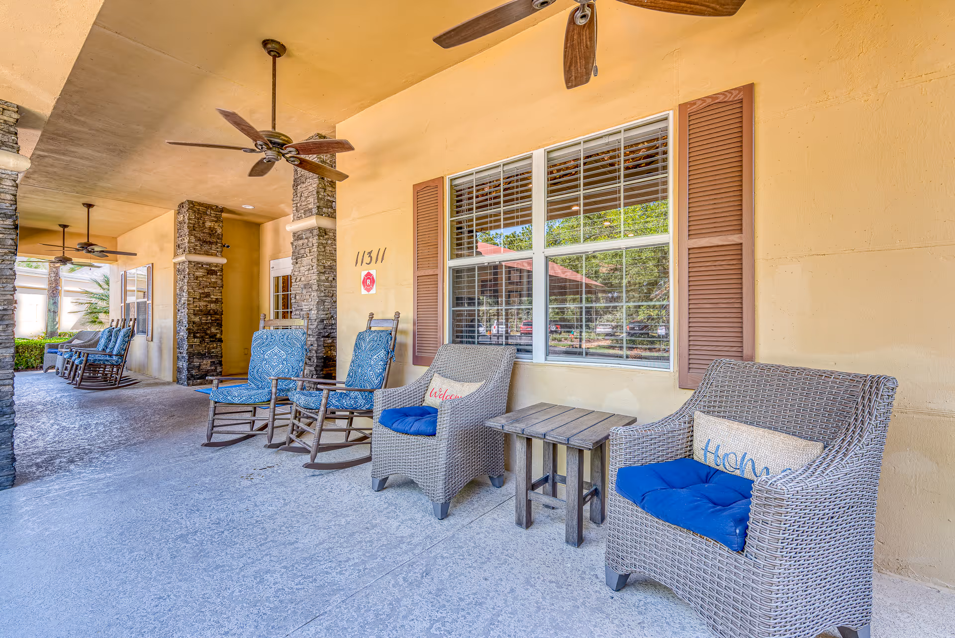 Covered outdoor patio area with wicker and wooden rocking chairs, some with blue cushions and pillows, ceiling fans, stone columns, and a window with brown shutters on a yellow wall.