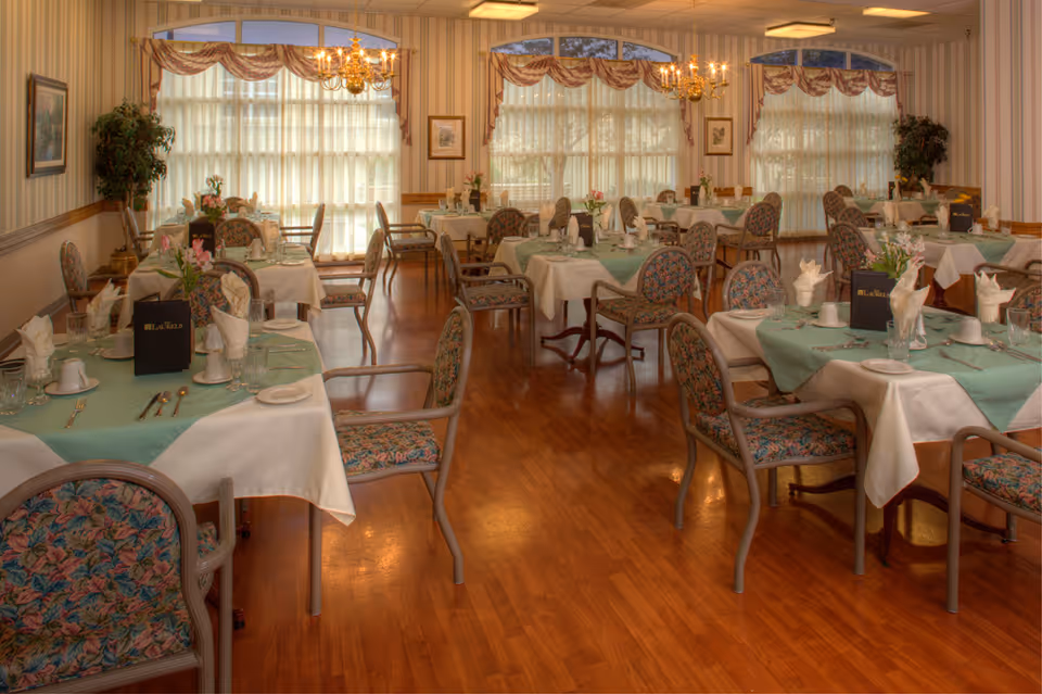 A warmly lit dining room with multiple tables covered in white and green tablecloths, each set with napkins, plates, cups, and silverware. The chairs have floral upholstery, and large windows with sheer curtains and valances allow natural light to fill the room. Two chandeliers hang from the ceiling, and there are framed pictures and potted plants along the walls.