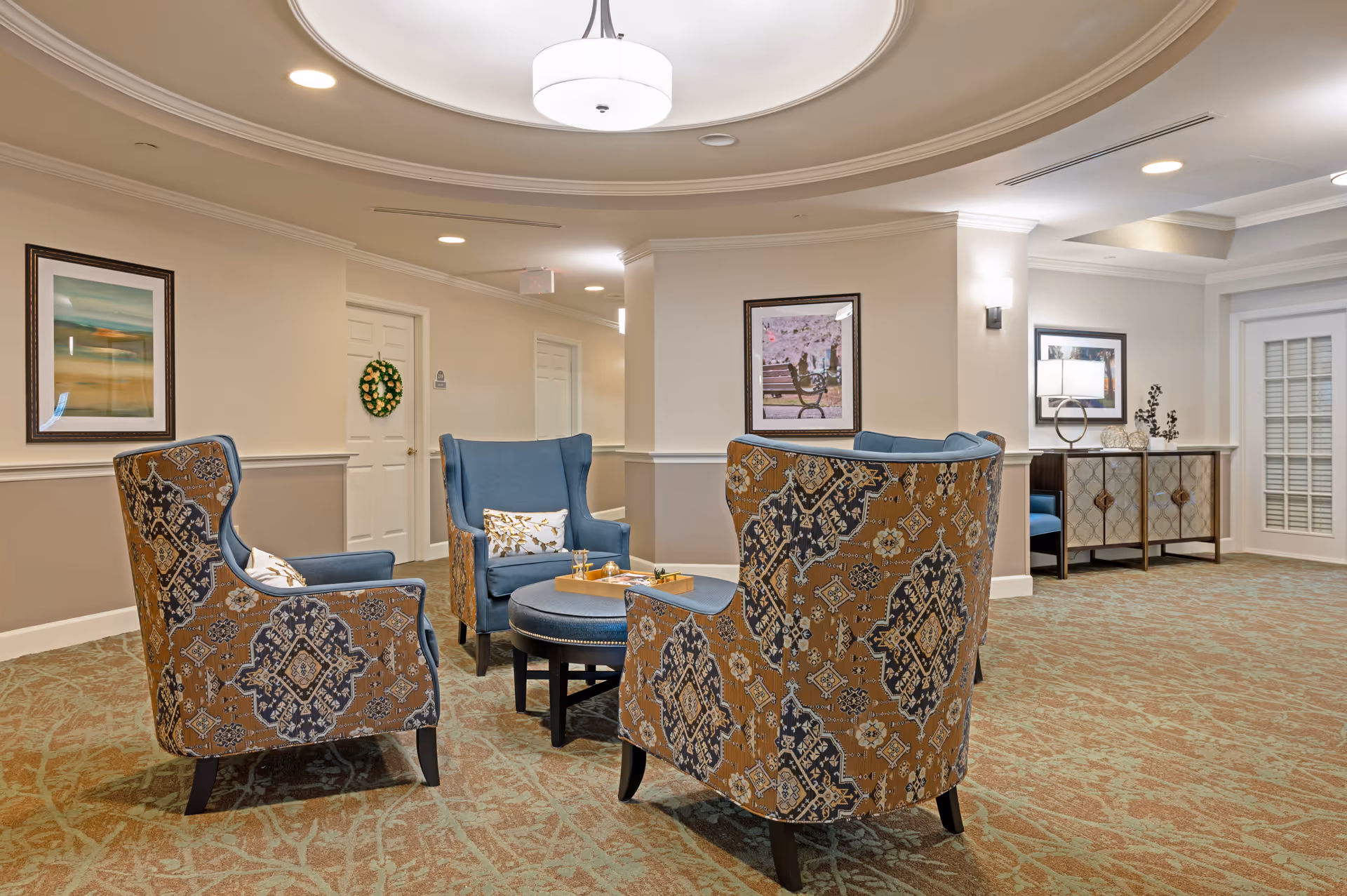A cozy seating area in a senior living facility with four upholstered armchairs arranged around a round ottoman. The chairs have a mix of blue fabric and patterned upholstery. The room features beige walls with white trim, framed artwork, a ceiling light fixture, and a sideboard with decorative items. The carpet has a green and brown pattern.