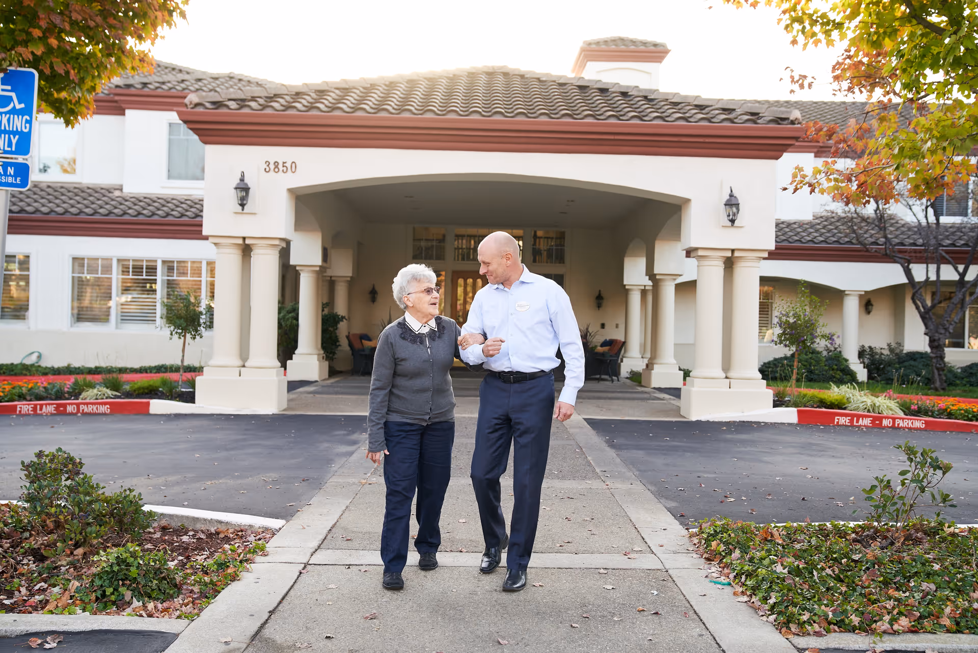 An elderly woman and a man walking arm in arm on a sidewalk in front of a building entrance with columns and a tiled roof. The building has the number 3850 above the entrance and there are trees with autumn leaves on either side of the walkway.