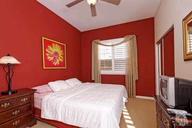 A bedroom with a red accent wall behind a bed with white bedding. There is a wooden nightstand with a lamp on the left side of the bed, a window with beige curtains, and a dresser with a TV on the right side. A ceiling fan is visible above.