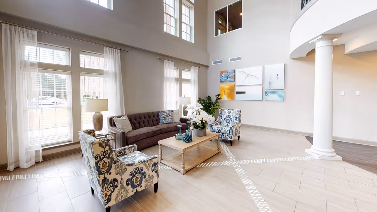 A bright and spacious living room area with high ceilings and large windows letting in natural light. The room features a dark gray tufted sofa, two patterned armchairs, a wooden coffee table with decorative items, two table lamps, and several pieces of artwork on the wall. There is a white column and light-colored tiled flooring with a decorative pattern.
