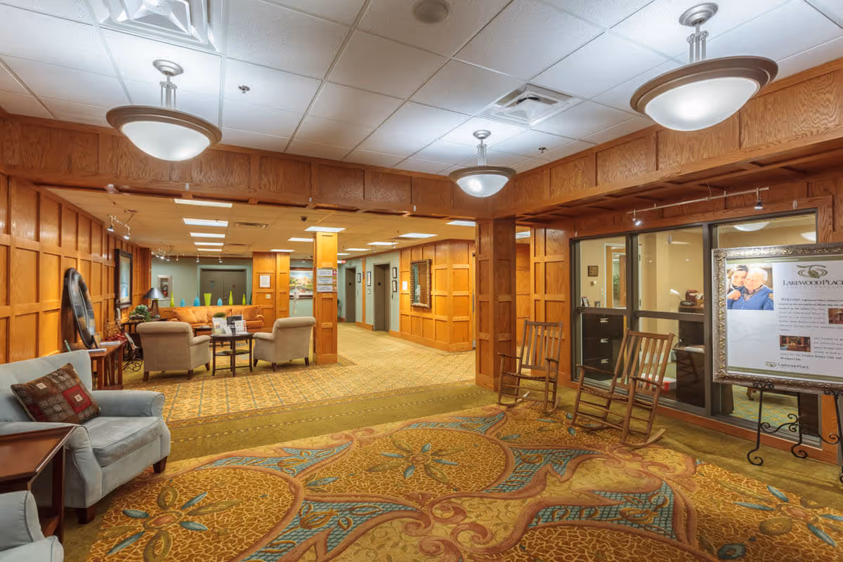Interior view of a senior living facility lobby area with wood-paneled walls, patterned carpet, comfortable seating including armchairs and rocking chairs, and ceiling lights. There is a framed informational board on the right side near glass windows and a hallway leading to elevators in the background.