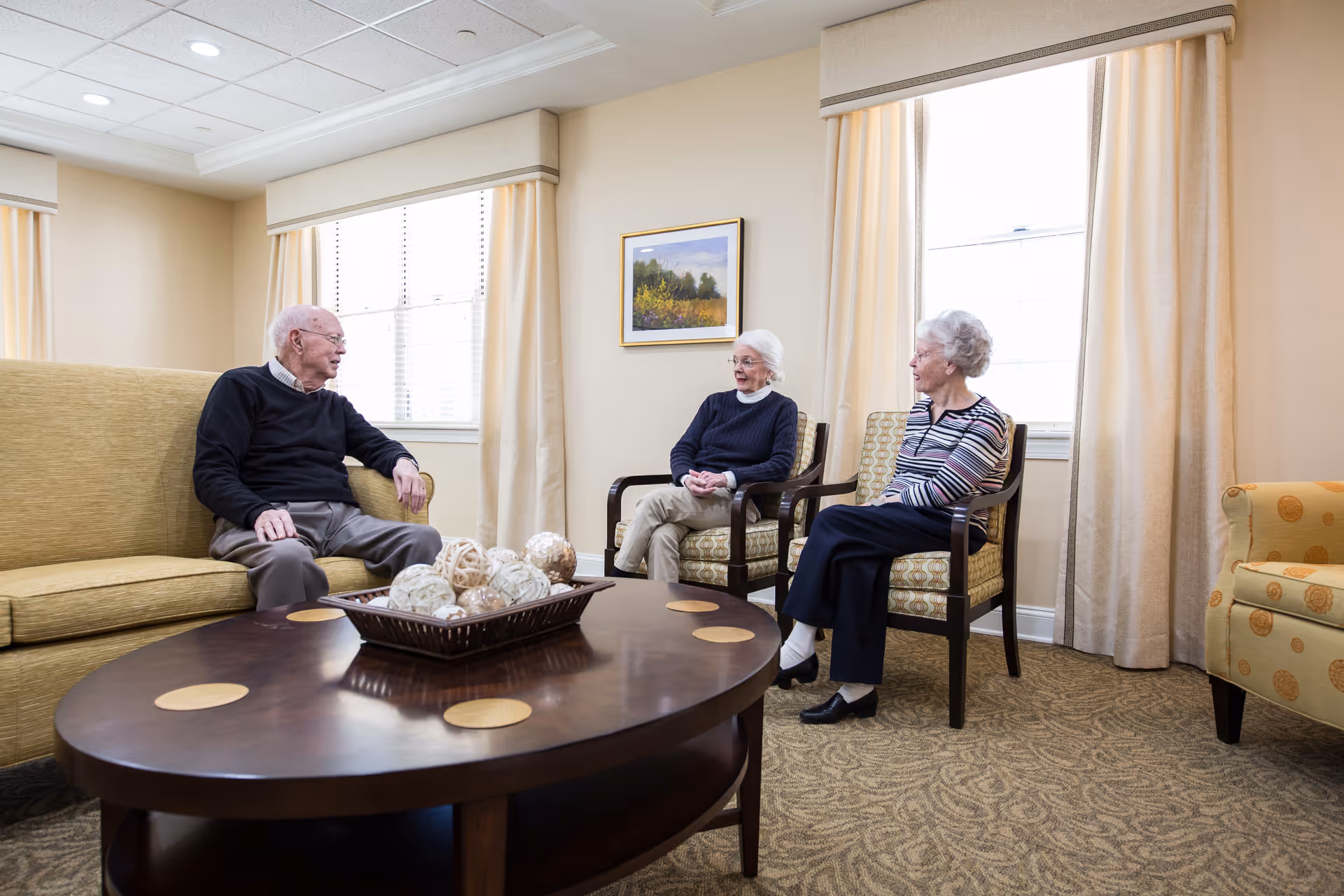 Three elderly people sitting and talking in a well-lit living room with beige walls, large windows with curtains, a wooden coffee table with decorative balls, and comfortable upholstered chairs and sofa.