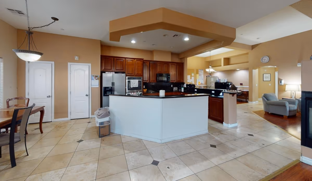 Interior view of a kitchen area in a senior living facility with beige walls, tiled floor, and wooden cabinets. The kitchen features a large white island with a black countertop, stainless steel refrigerator, oven, and microwave. To the left, there is a dining table with chairs and a hanging light fixture. In the background, there is a seating area with an armchair and a lamp.