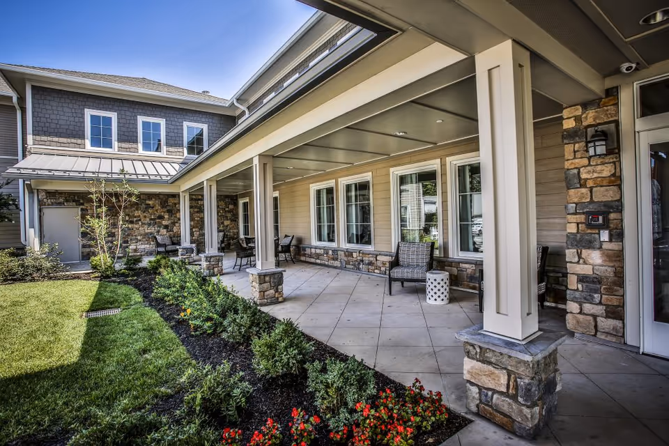 Covered outdoor patio area at Senior Living Residences Marlboro with stone pillars, several chairs, small tables, and a landscaped garden with green grass and flowering plants.