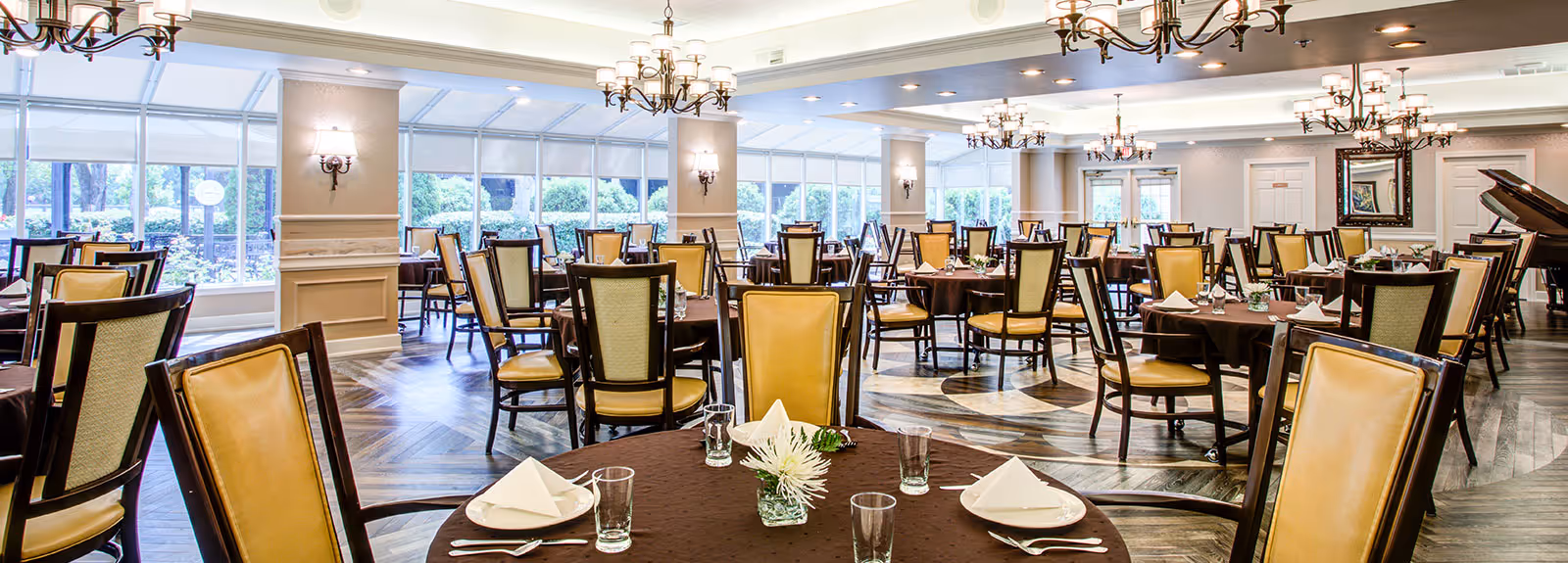 Spacious dining room with round tables set for service, high-backed chairs, chandeliers and large windows overlooking greenery.