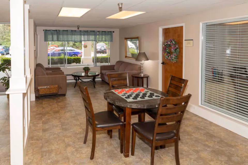 A cozy common area in a senior living facility featuring a round table with four wooden chairs and a checkers board on top. In the background, there are two sofas, a glass coffee table with a small plant, a side table with a lamp, a wall mirror, and a door decorated with a floral wreath. Large windows with green valances let in natural light and show parked cars outside.