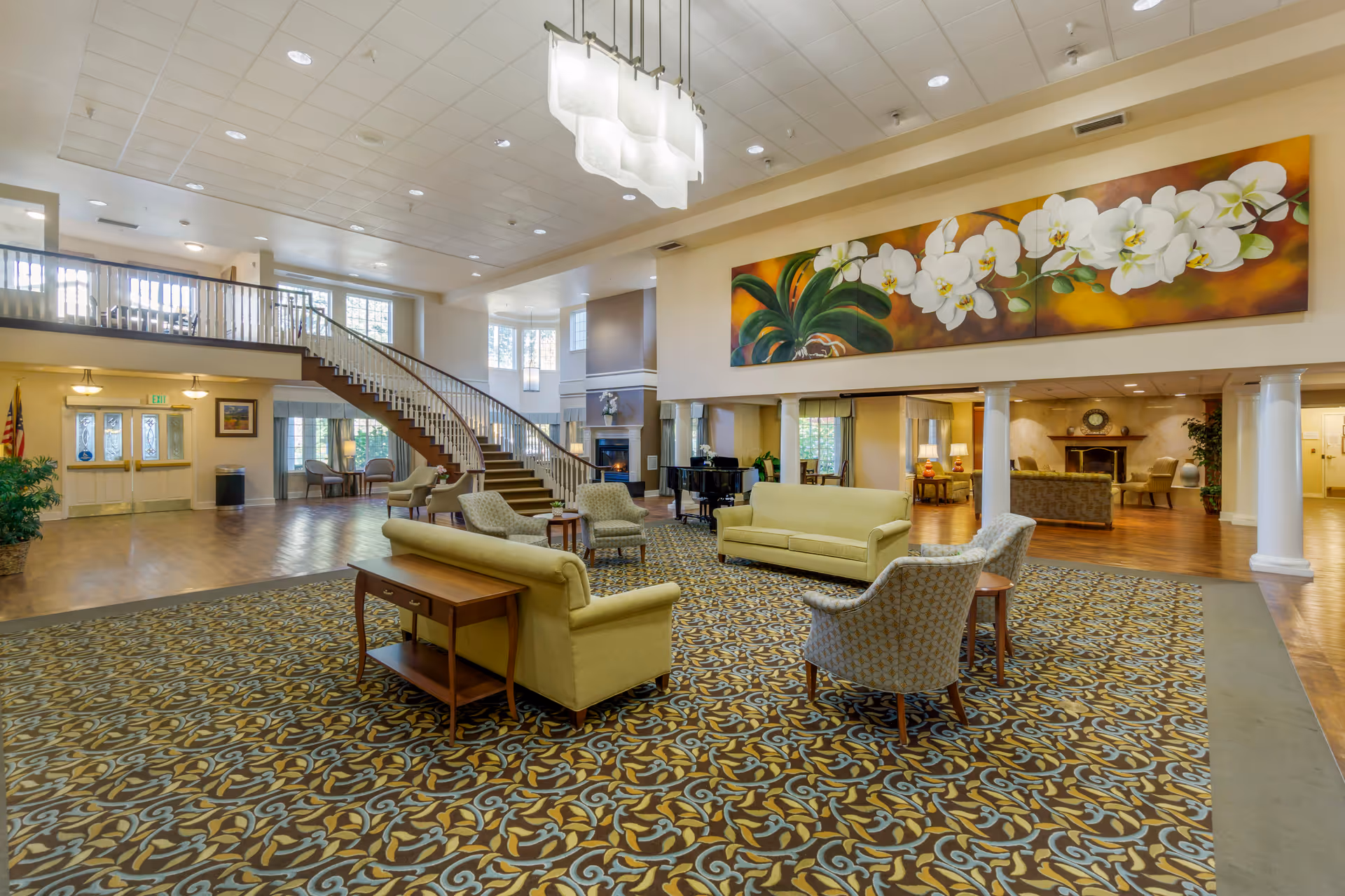 Spacious and well-lit senior living facility common area with patterned carpet, multiple seating arrangements including sofas and armchairs, a grand piano, a staircase leading to an upper level, and a large floral painting on the wall.