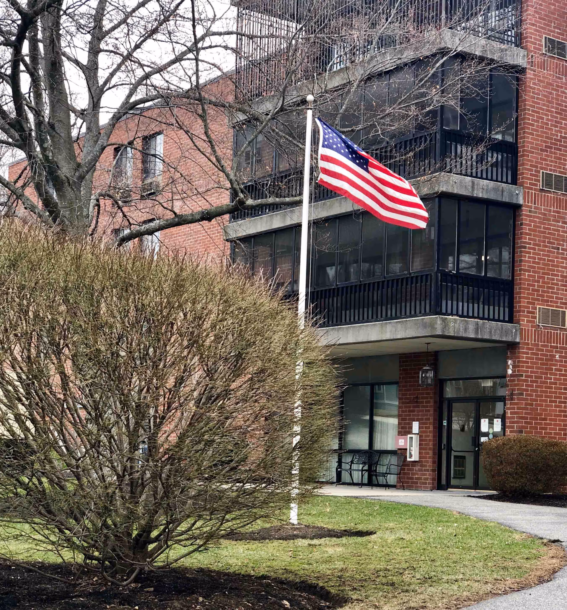 Exterior view of a brick building with a glass-enclosed balcony and an American flag on a flagpole in front. There are leafless trees and bushes around the building, and a pathway leading to the entrance.