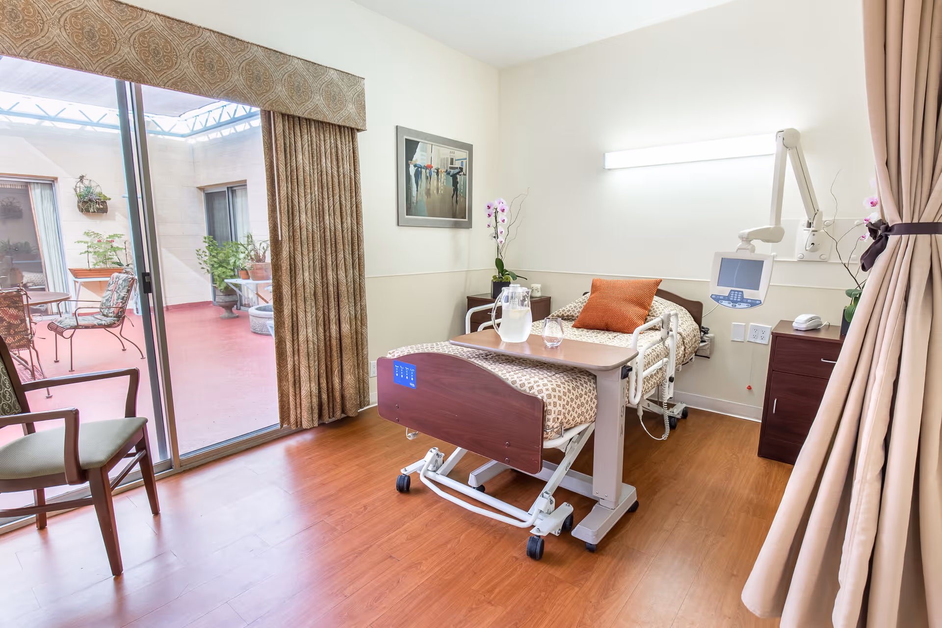A bright senior care patient room with a hospital bed, bedside table, sliding glass door to a patio, and chairs.