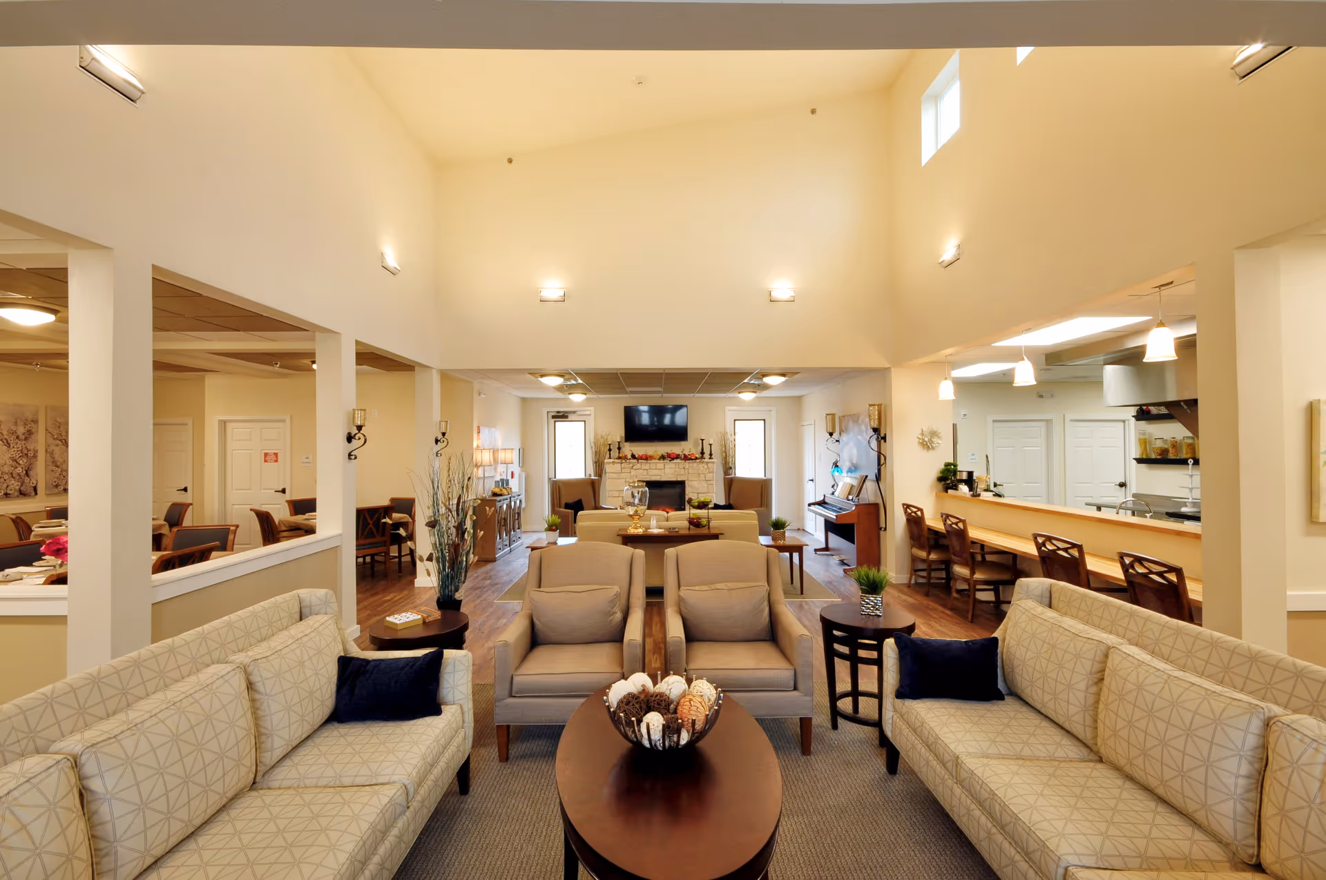 A spacious senior living facility common area with beige walls and high ceilings. The room features two beige patterned sofas with dark blue pillows, two beige armchairs, and a dark wooden oval coffee table with decorative items. In the background, there is a fireplace with a TV mounted above it, a piano to the right, and a counter with bar stools on the right side. The room is well-lit with wall sconces and pendant lights.