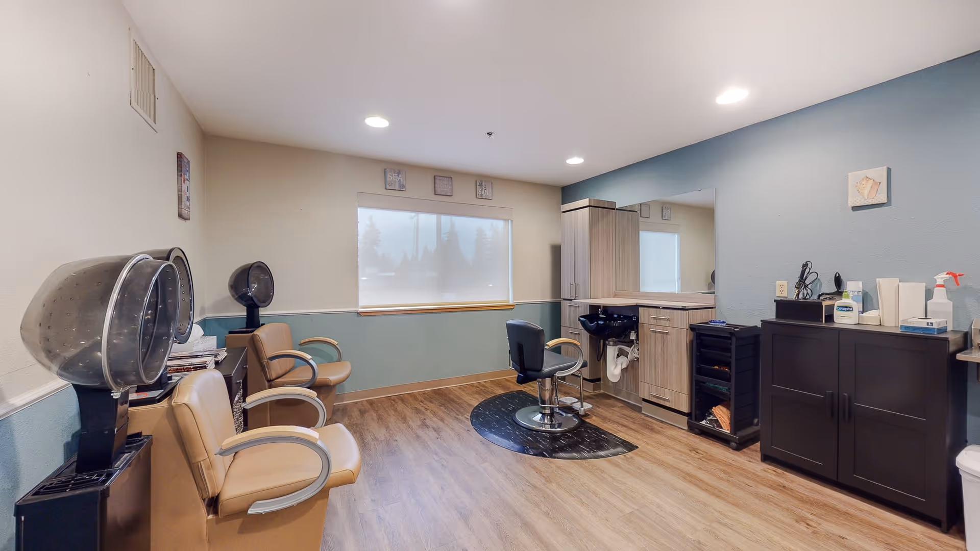 Interior view of a hair salon area in a senior living facility with beige salon chairs, hair dryers, a black salon chair in front of a mirror, wooden cabinets, and various hair care products on counters. The walls are painted light blue and white, and there is a window with a shade partially drawn.