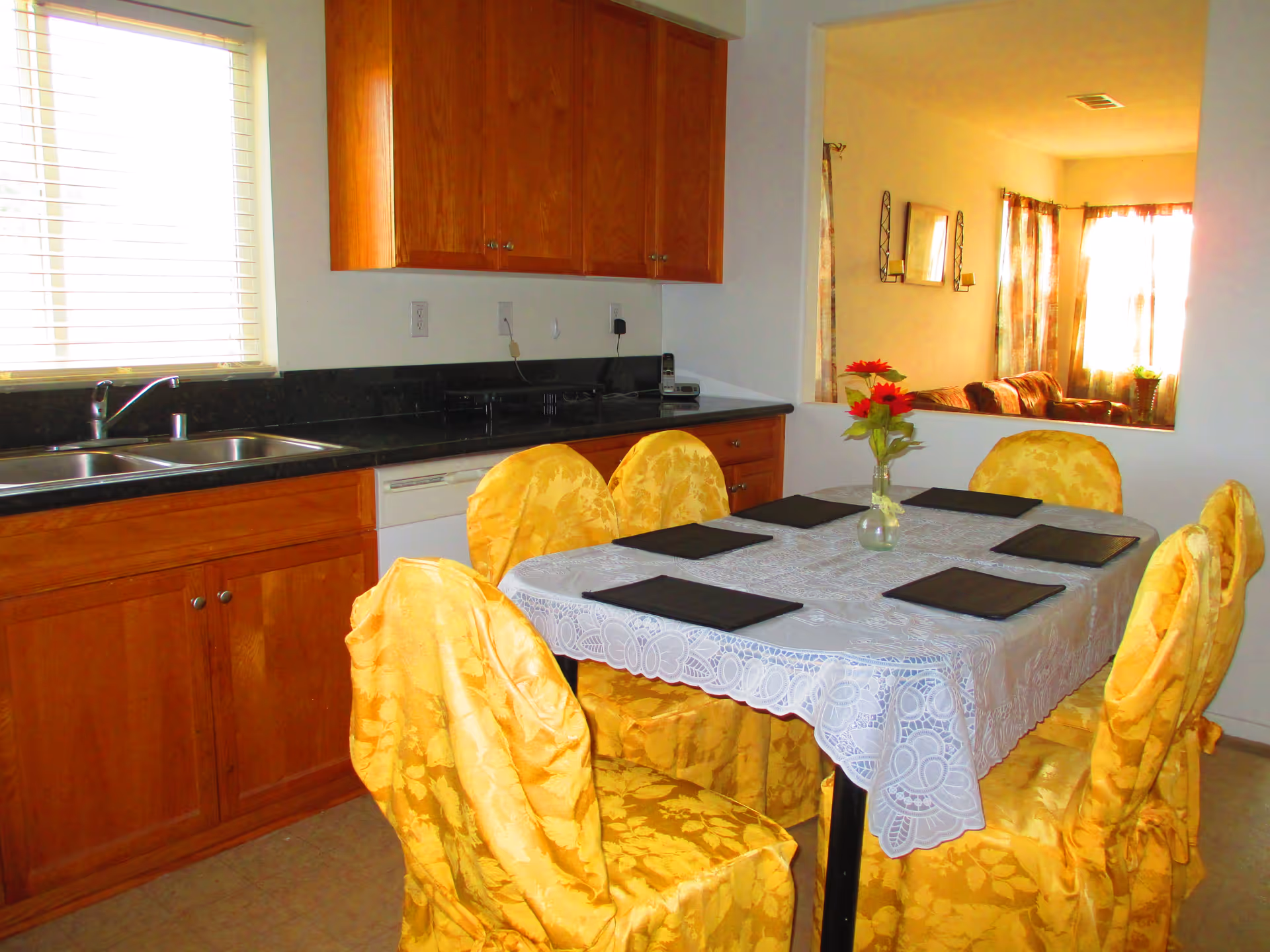 A dining area with a rectangular table covered with a white lace tablecloth and six chairs draped in yellow floral fabric covers. The table has six black placemats and a small vase with red flowers. The dining area is adjacent to a kitchen with wooden cabinets, a black countertop, a double sink, and a window with blinds. In the background, a living room with a sofa and curtains is visible through a large interior window opening.