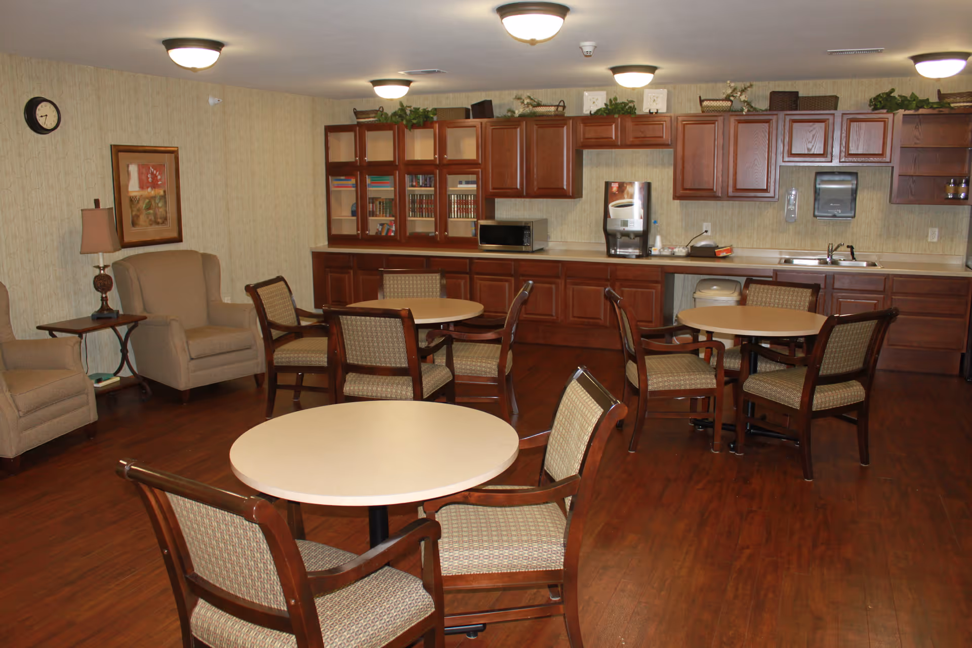 A common area in a senior living facility featuring round tables with chairs, a kitchenette with wooden cabinets, a microwave, a coffee machine, and a sink. There are also two armchairs with a side table and a lamp, a wall clock, and framed artwork on the wall.