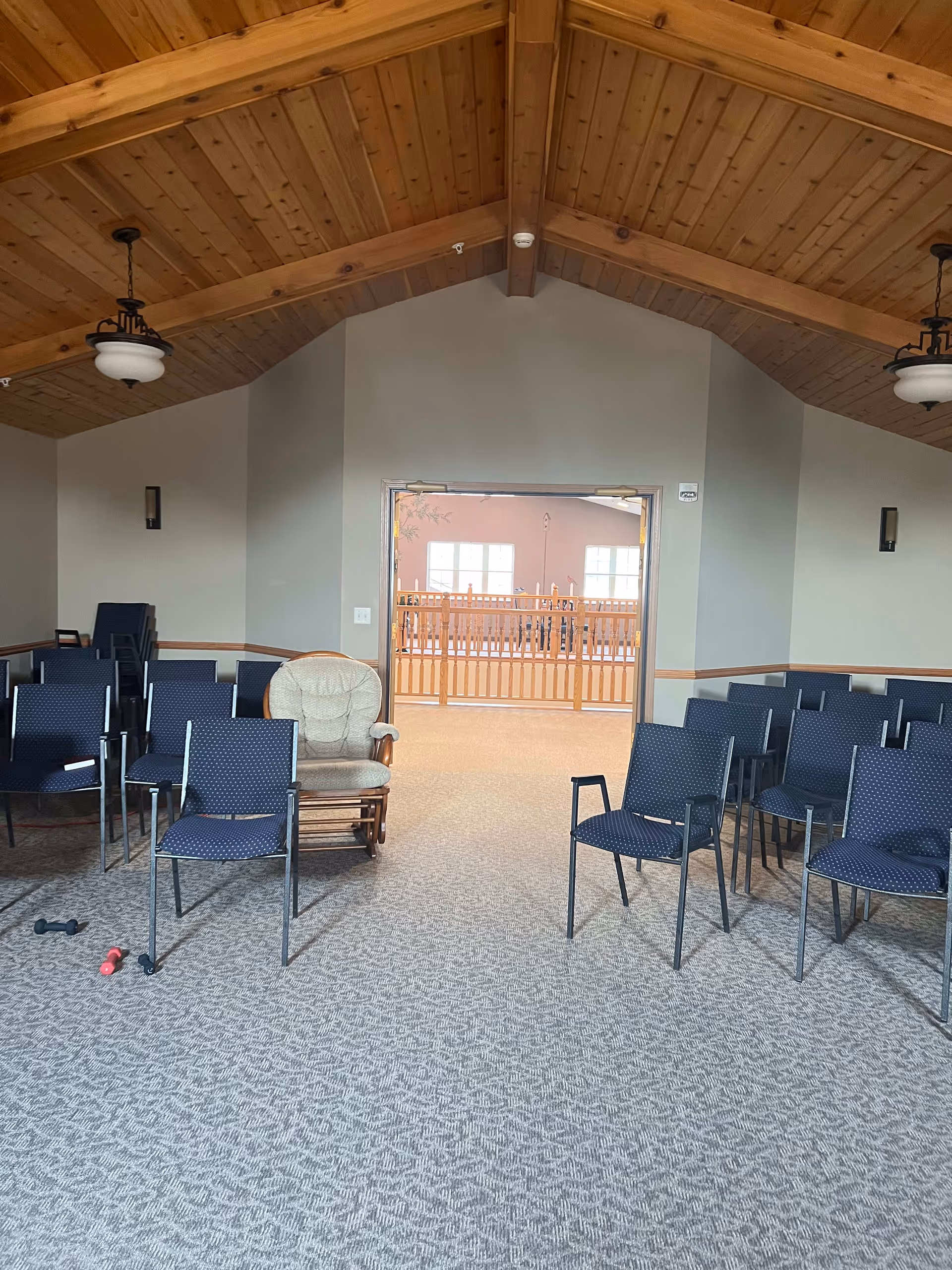 Interior room with a high wooden ceiling and carpeted floor, arranged with multiple blue chairs and one cushioned rocking chair. The room has two hanging light fixtures and wall sconces. An open doorway leads to another area with wooden railings and large windows.
