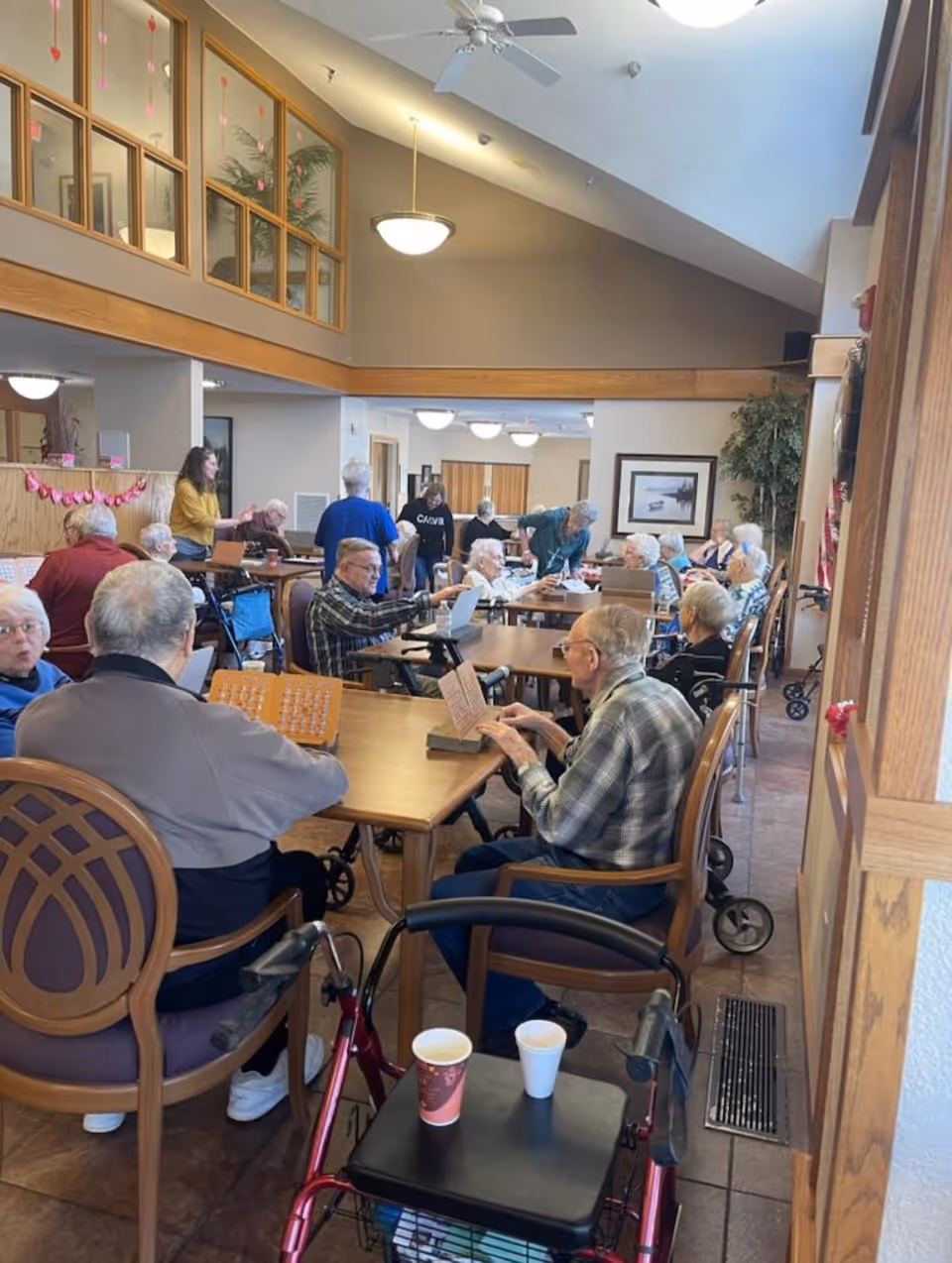 A group of elderly people sitting around tables in a spacious, well-lit common area of a senior living facility. Some are engaged in activities like reading or playing games, while others are conversing. The room has high ceilings, ceiling fans, and large windows. There are walkers and cups on the tables, and a few staff members are assisting residents.