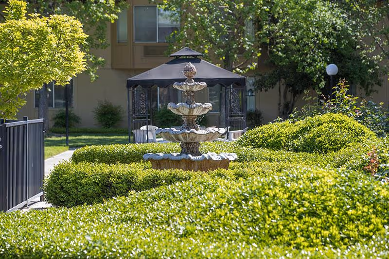 A multi-tiered stone fountain surrounded by neatly trimmed green bushes in an outdoor garden area. In the background, there is a gazebo with a dark roof and some chairs underneath, along with trees and a building facade.