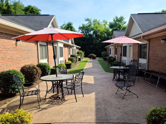Outdoor patio area between two brick buildings with round metal tables, black metal chairs, and red umbrellas providing shade. The area is surrounded by green bushes and trees in the background under a clear blue sky.