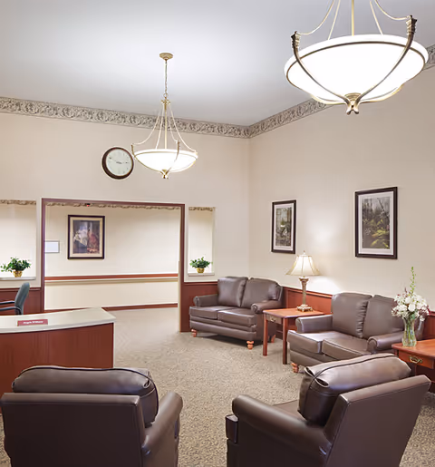 A well-lit common area with beige walls and carpeted floor featuring two brown leather sofas, two brown leather armchairs, wooden side tables with a lamp and a vase of flowers, framed pictures on the walls, two hanging ceiling lights, and a clock above an open doorway.