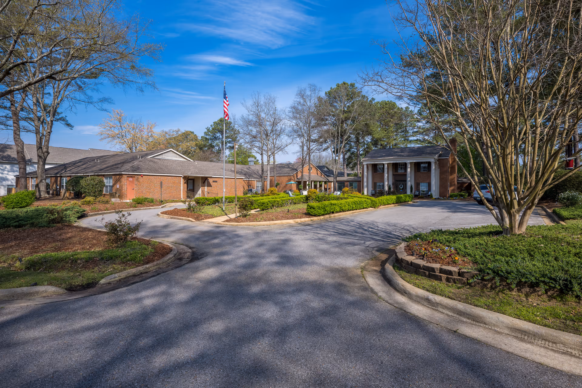 Exterior view of Pine Valley Retirement Community showing a circular driveway with landscaped greenery and an American flag in the center. The building is a single-story brick structure with a covered entrance supported by white columns. Trees with sparse leaves surround the area under a clear blue sky.