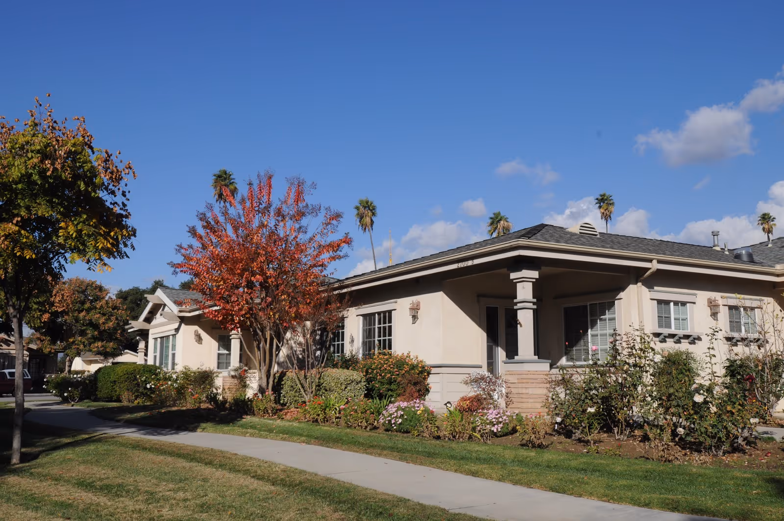Exterior view of a single-story residential building with beige walls and a gray roof, surrounded by well-maintained landscaping including green bushes, flowering plants, and trees with autumn foliage. A concrete sidewalk runs alongside the building under a clear blue sky with a few clouds and palm trees in the background.