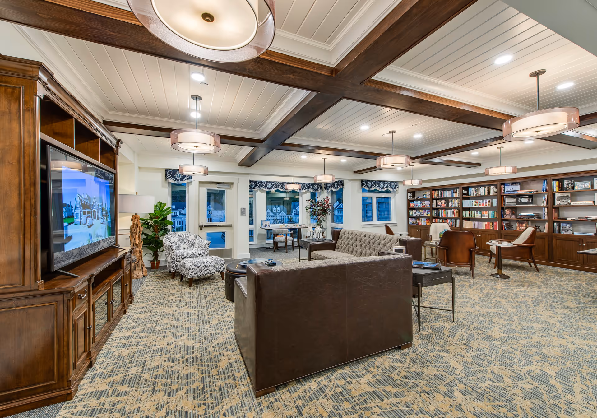 A spacious and well-lit living room area with a patterned carpet, wooden ceiling beams, and multiple pendant lights. The room features a large wooden entertainment center with a flat-screen TV, a gray tufted sofa, a dark brown leather sofa, patterned armchairs, and several wooden chairs. There is a large bookshelf filled with books and games along one wall, and windows with blue and white valances letting in natural light.