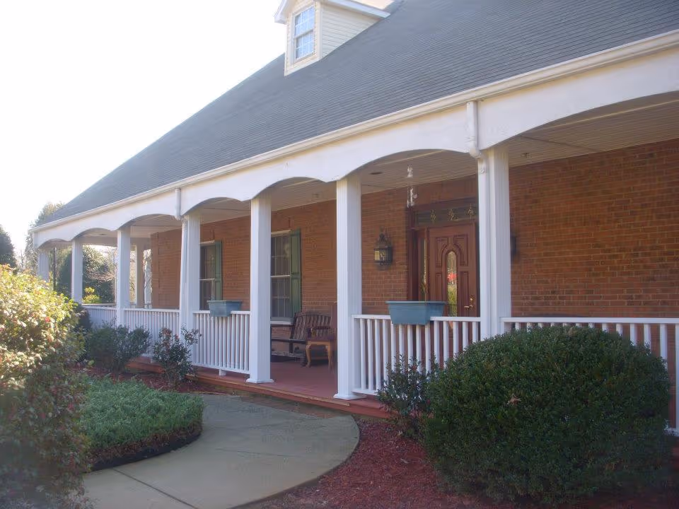 Front porch of a brick building with white columns and railing, a wooden bench, two green window shutters, and a red door. There are bushes and plants along the walkway leading to the porch.