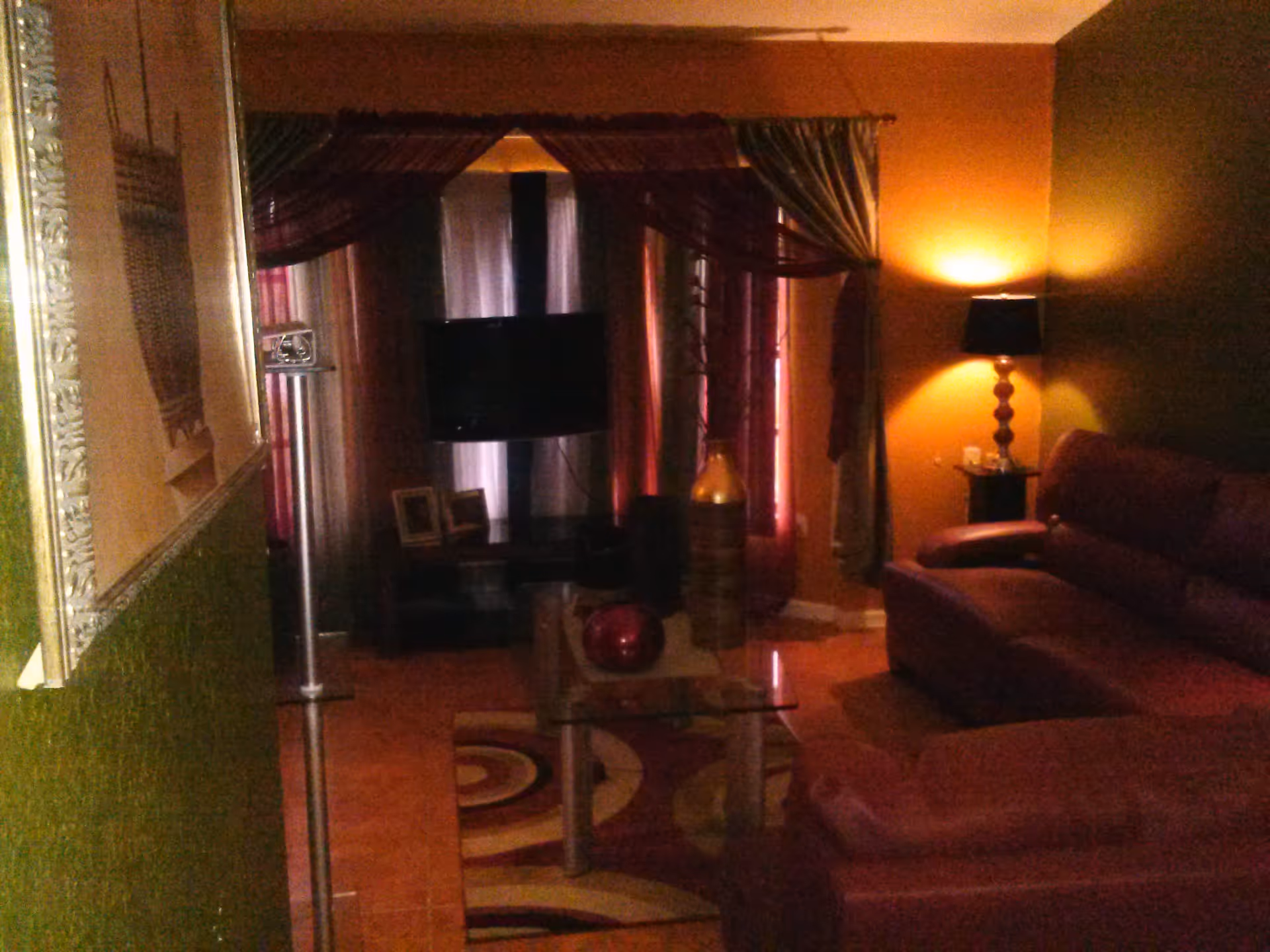 Dimly lit living room with maroon couches, a glass coffee table with decorative items, a floor lamp with a black shade, and a TV mounted in front of windows covered with dark curtains.