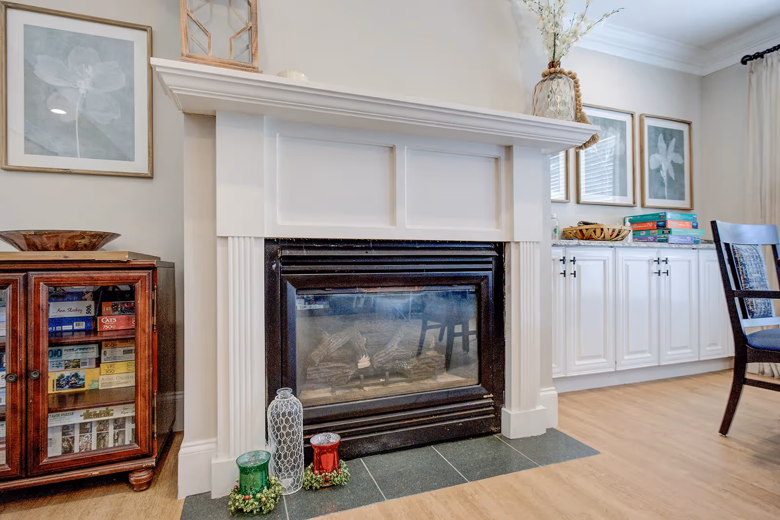 A cozy living room area featuring a white fireplace with a black insert. On the floor in front of the fireplace are decorative glass candle holders in green, white, and red. To the left is a wooden cabinet with glass doors displaying board games and puzzles. On the right side, there is a white cabinet with black handles topped with a basket and several board games. Above the cabinet are three framed floral artworks. A dark wooden chair with a cushion is partially visible on the right side. The room has light-colored walls and wood flooring.