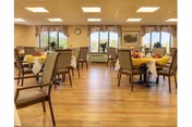 A spacious dining room with multiple tables covered in yellow tablecloths and set with napkins and utensils. The room has wooden flooring, large windows with floral valances, and beige walls decorated with framed artwork. Several chairs are arranged around each table.
