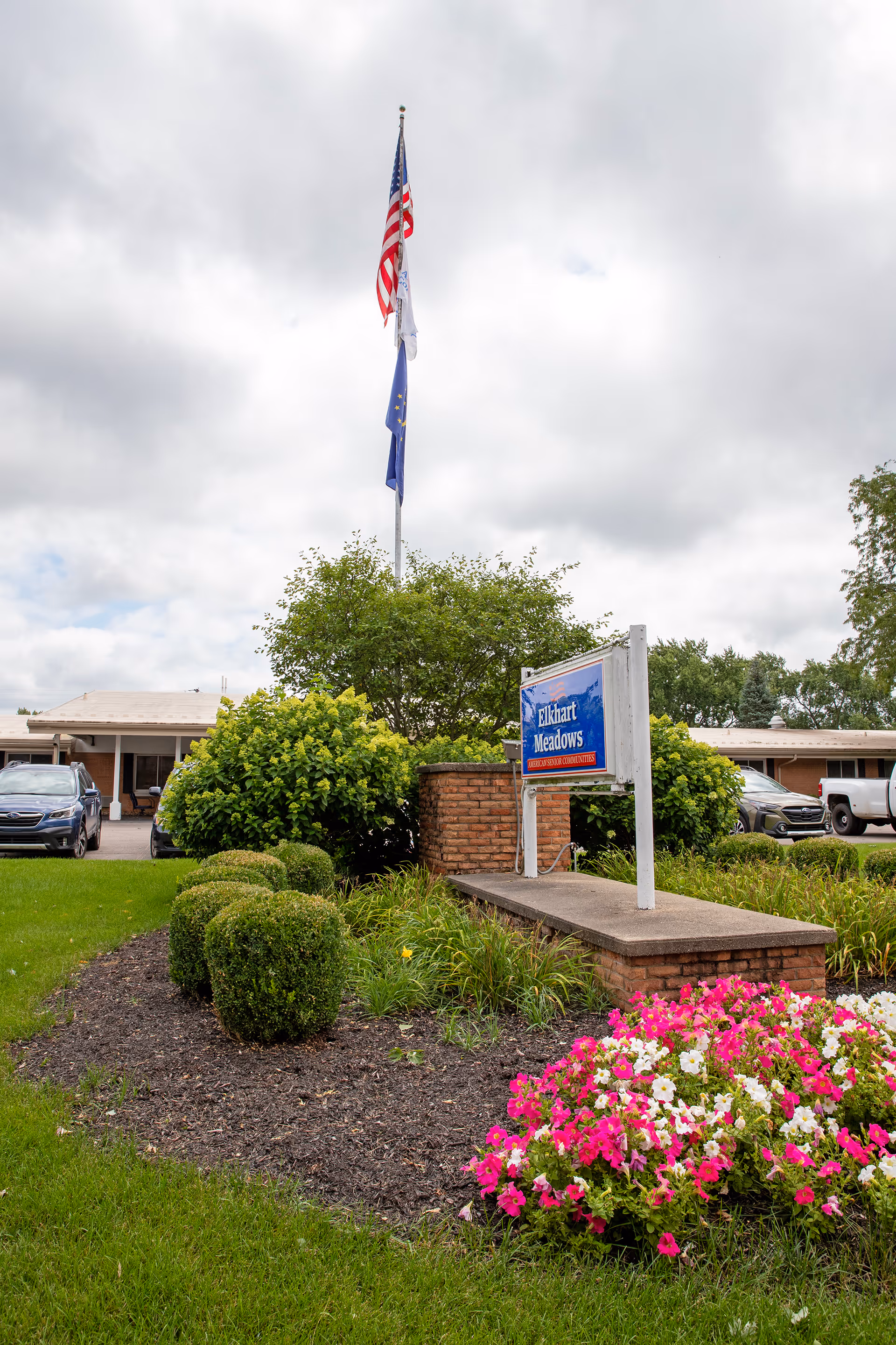 Entrance sign for Elkhart Meadows with a flagpole and landscaped flowerbeds in front of the building.