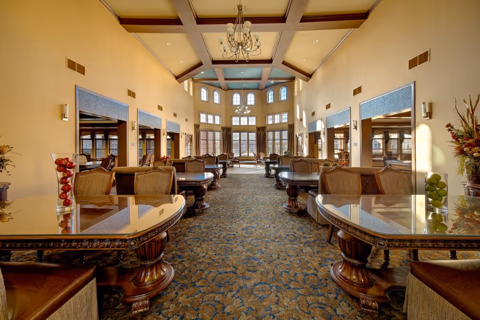 A spacious and elegant dining room with high ceilings and large windows allowing natural light to fill the space. The room features multiple round wooden tables with cushioned chairs arranged neatly along a patterned carpet. Decorative chandeliers hang from the ceiling, and vases with fruit and floral arrangements are placed on the tables and side surfaces.