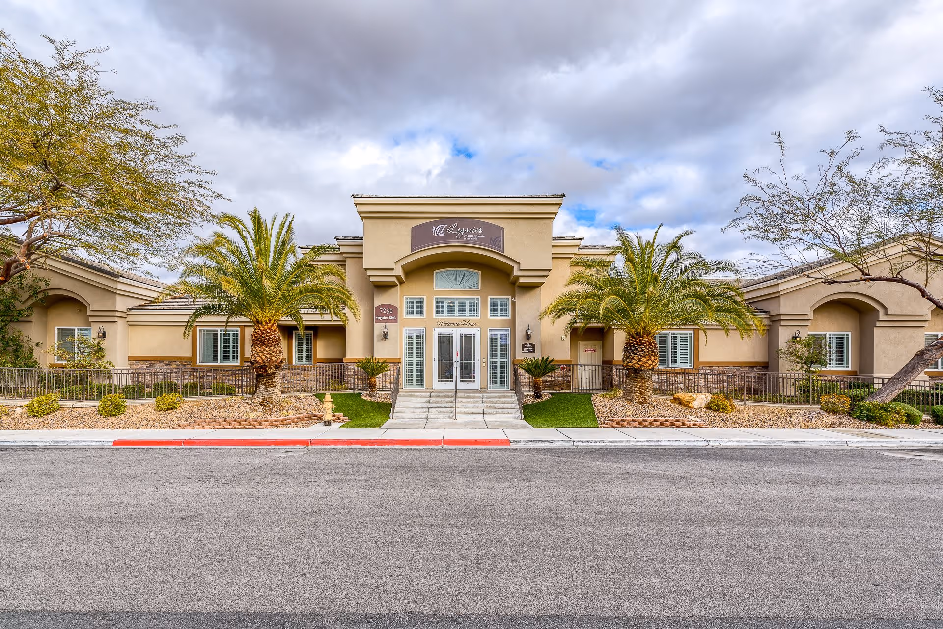 Front exterior view of a single-story senior living facility building with beige walls, large windows, and a central entrance with glass doors. The entrance is flanked by palm trees and landscaped with rocks and small bushes. The sky is partly cloudy.