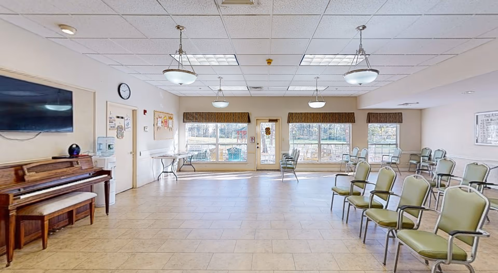A spacious community room with tiled floors and beige walls featuring multiple green cushioned chairs arranged in rows. There is a wooden piano with a bench on the left side under a mounted flat-screen TV. The room has large windows and a glass door at the far end letting in natural light. Ceiling lights hang from a white tiled ceiling. A water cooler and bulletin boards are visible near the door on the left wall.