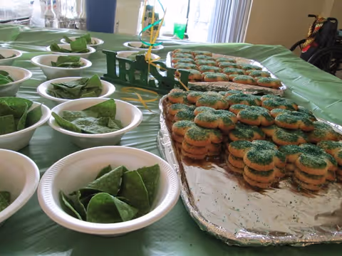 A table covered with a green tablecloth displaying multiple white bowls filled with green tortilla chips and trays of stacked cookies decorated with green sugar sprinkles. A small decorative sign with the word 'Irish' is also on the table.