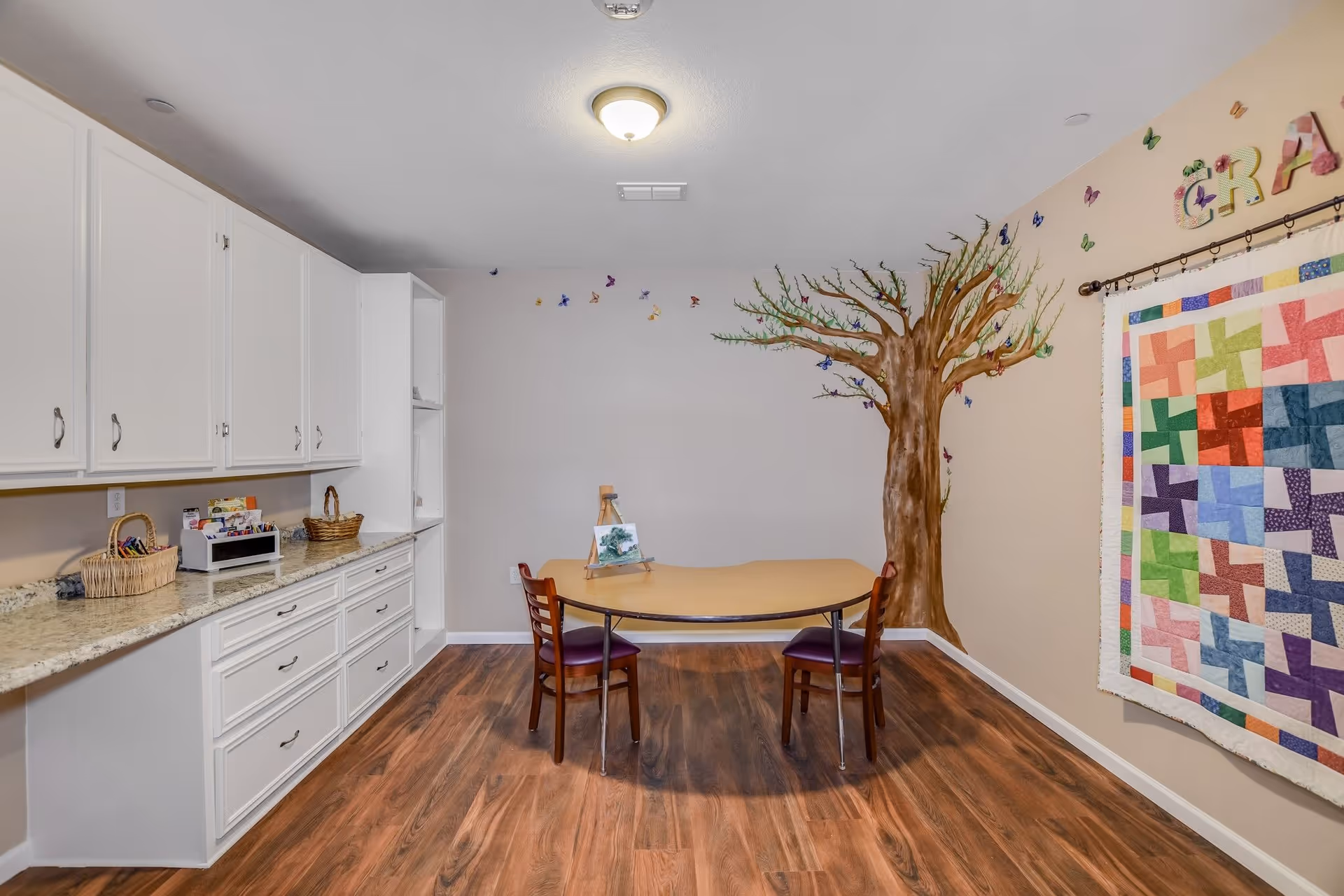 A craft room with a wooden table and two chairs on a wood floor. The walls are decorated with a painted tree with butterflies and a colorful quilt hanging on the right wall. White cabinets with drawers and baskets are on the left side of the room.