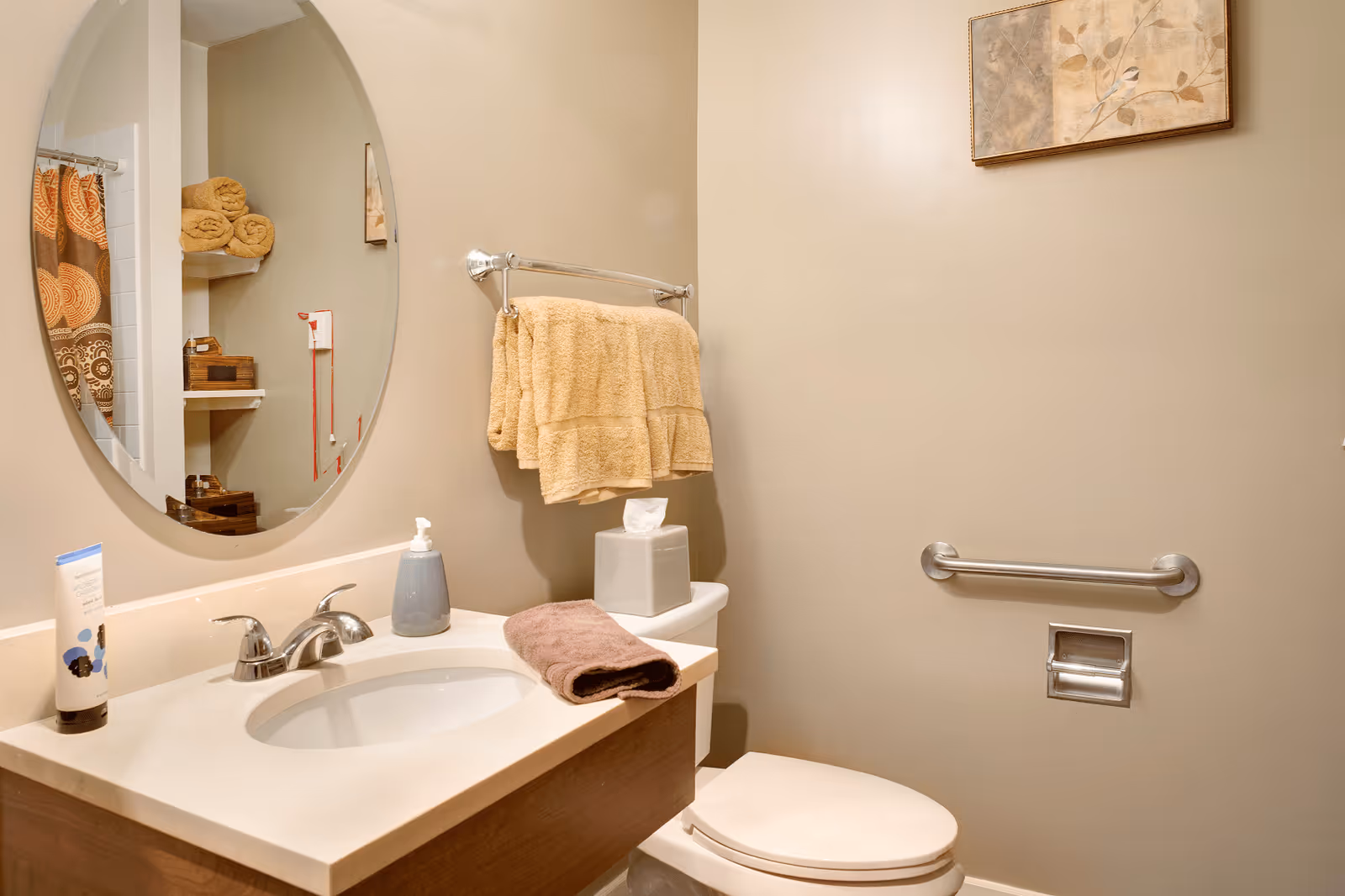 A clean and well-lit bathroom featuring a sink with a silver faucet, a soap dispenser, a brown towel on the counter, a toilet with a tissue box on top, beige towels hanging on a towel rack, a grab bar next to the toilet, and a round mirror reflecting shelves with rolled towels and decorative items.