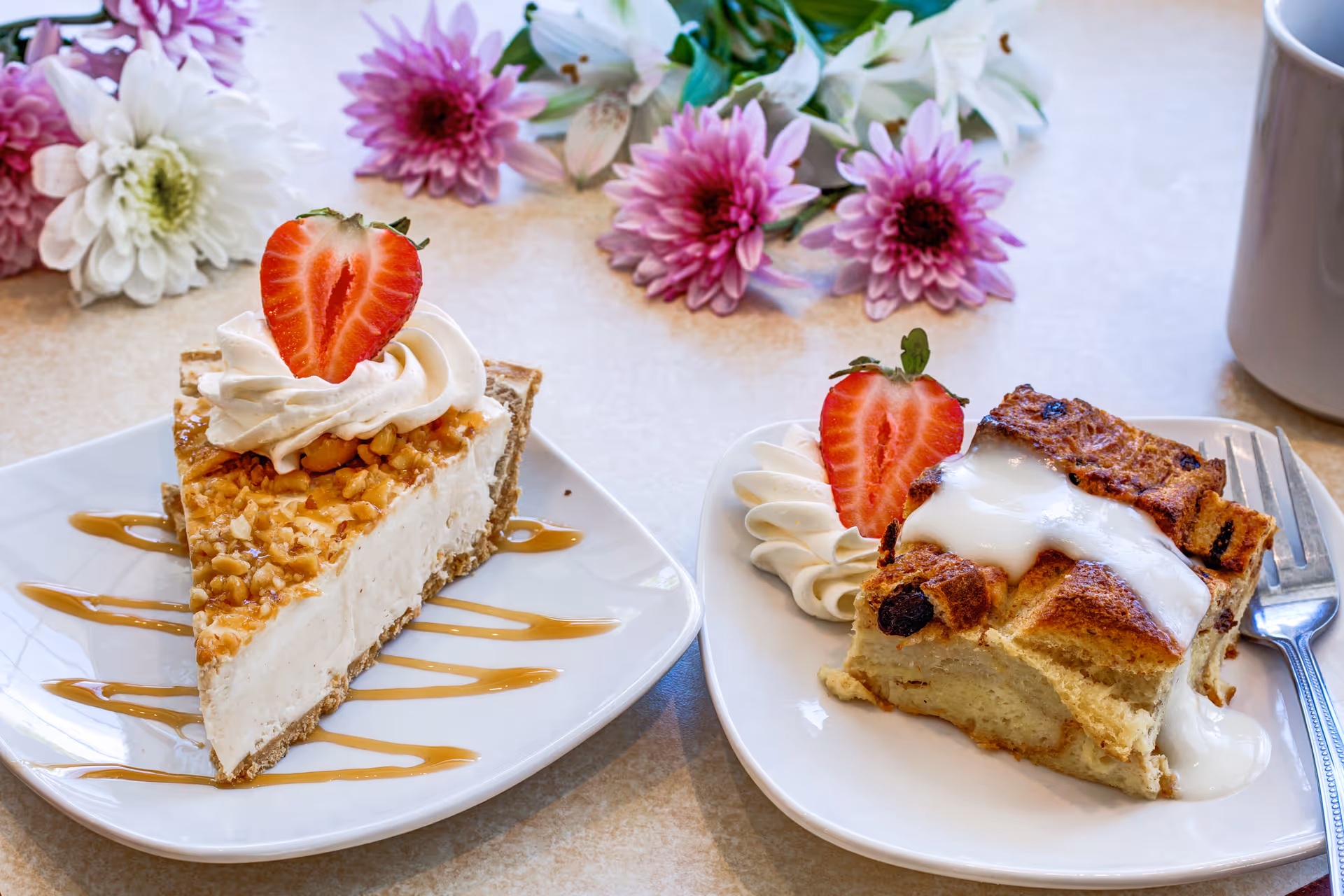Two dessert plates on a table with a background of pink and white flowers. One plate has a slice of nut-topped cheesecake with whipped cream and a strawberry half on top, drizzled with caramel sauce. The other plate has a piece of bread pudding topped with white icing, whipped cream, and a strawberry half, with a fork beside it. A white mug is partially visible on the right side.