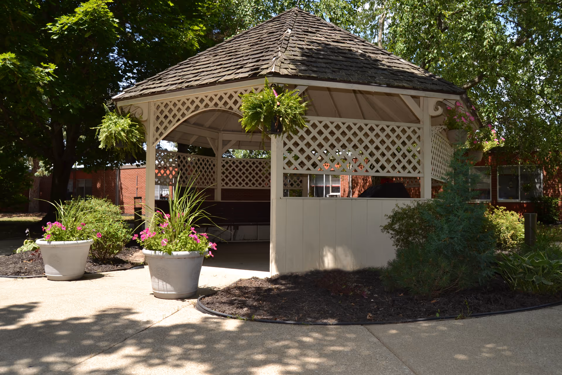 A white wooden gazebo with lattice trim sits in a landscaped courtyard with potted flowers and trees in front of a brick building.