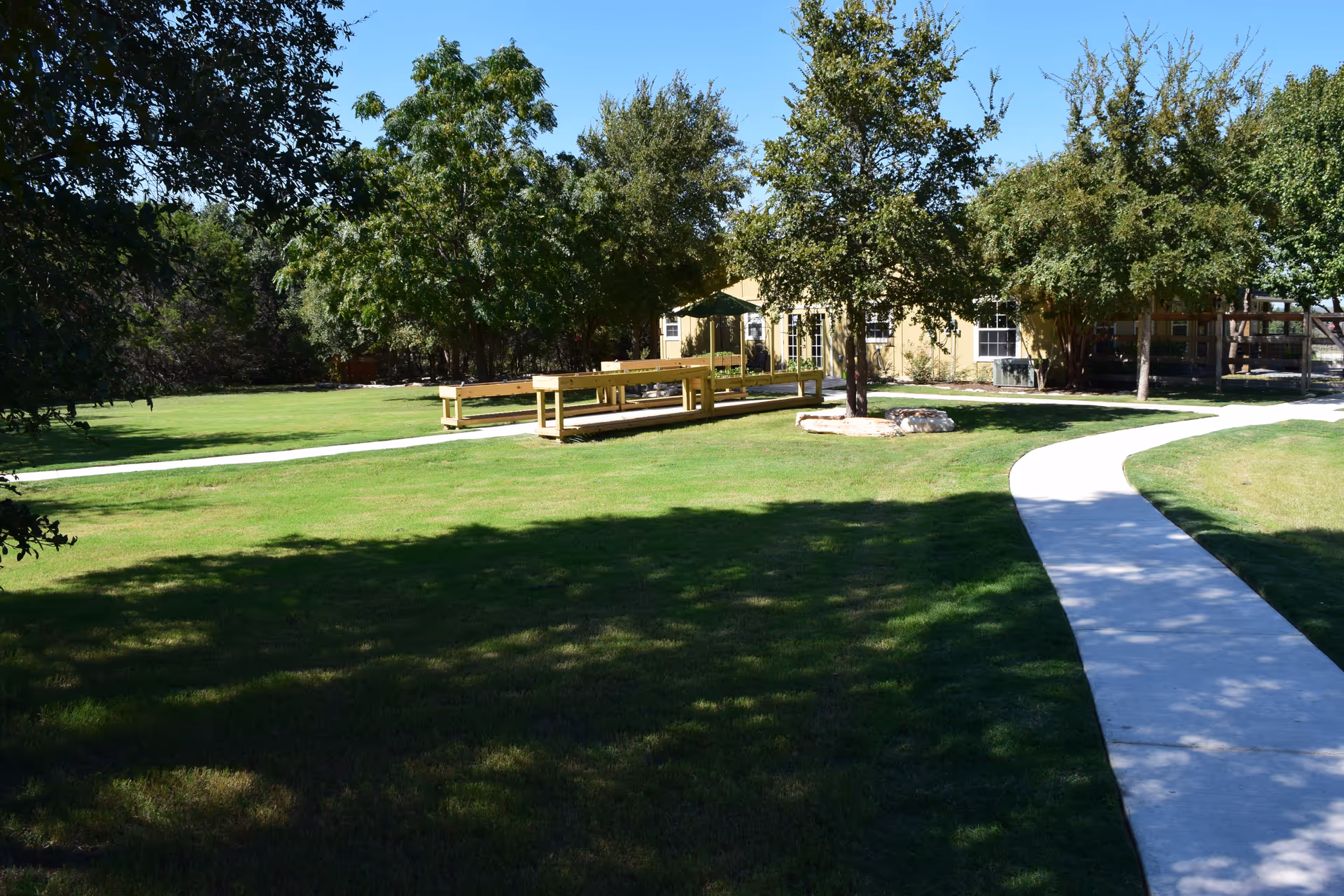 A sunny outdoor area with a curved concrete walkway leading through a green lawn with several trees. There is a wooden picnic table with benches under the shade of the trees, and a light-colored building is visible in the background.