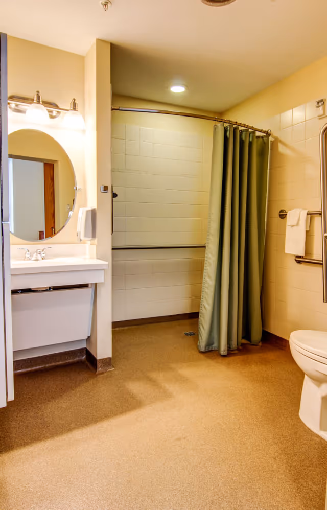 A clean and accessible bathroom with a white sink and mirror on the left, a shower area with a green curtain in the center, and a toilet on the right. The bathroom has beige walls and flooring, grab bars near the shower and toilet, and a towel hanging on the wall.