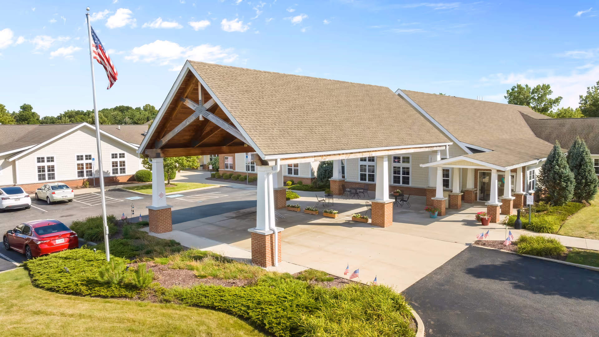 Exterior view of Hudson Springs Nursing & Rehab facility showing the main entrance with a covered drop-off area supported by white columns with brick bases. There is a flagpole with an American flag, several parked cars, and well-maintained landscaping with bushes and grass under a clear blue sky.